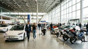 Vue réaliste du Parc Expo de Tours pendant un salon automobile, avec un homme et une femme parmi des visiteurs observant des voitures électriques et des motos dans un hall lumineux.